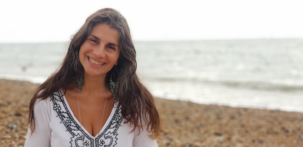 Smiling woman with long dark hair, wearing a white embroidered top, standing on a beach with a blurred ocean background.