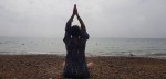 A person sitting cross-legged on a beach, practicing a spiritual or mindfulness pose with their hands raised above their head, against a cloudy sky and tranquil sea.